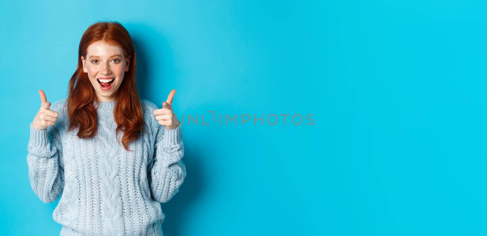 Cheerful teenage girl showing approval, make thumbs up and agree, liking something, standing over blue background.