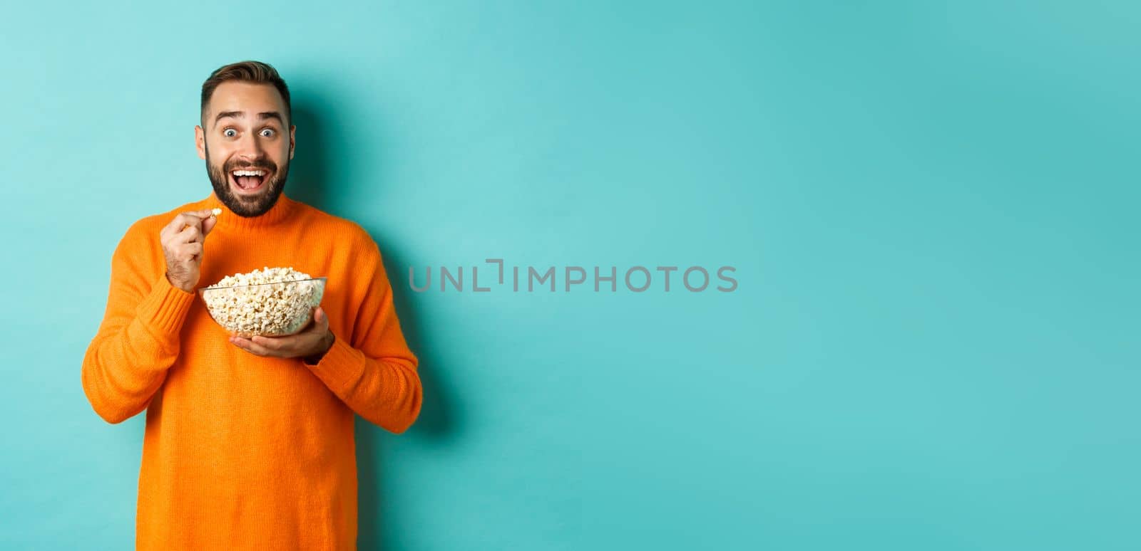 Excited young man watching interesting movie on tv screen, eating popcorn and looking amazed, blue background.