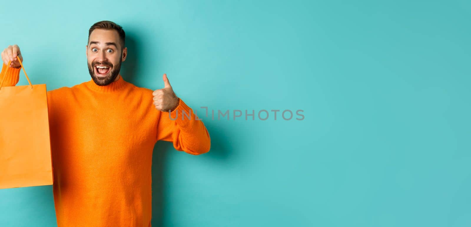 Happy man holding orange shopping bag and rejoicing from discount and celebrating, showing thumbs up and recommending, standing over blue background.