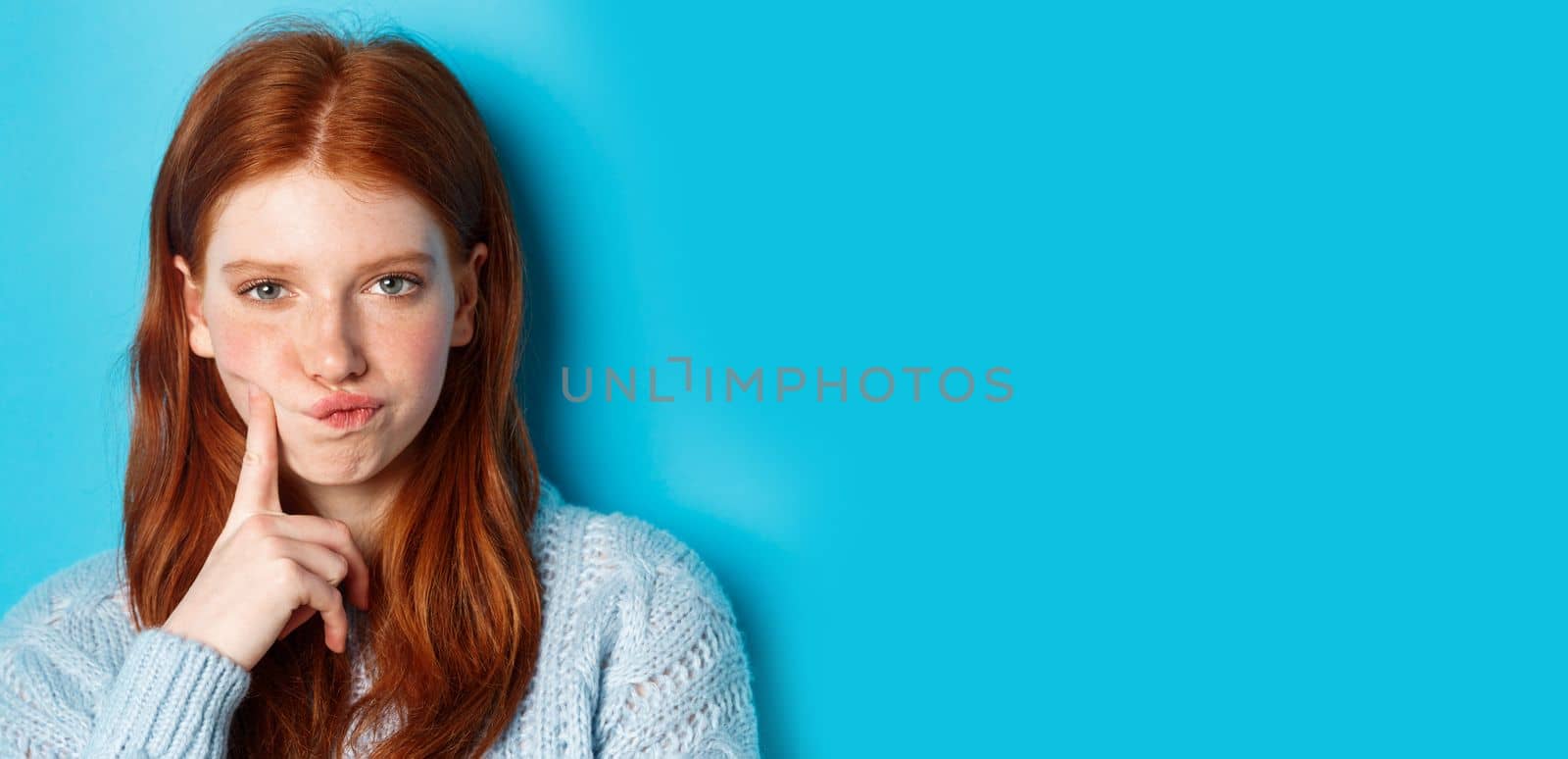 Puzzled redhead girl looking suspicious at camera, thinking or solving problem, standing against blue background.