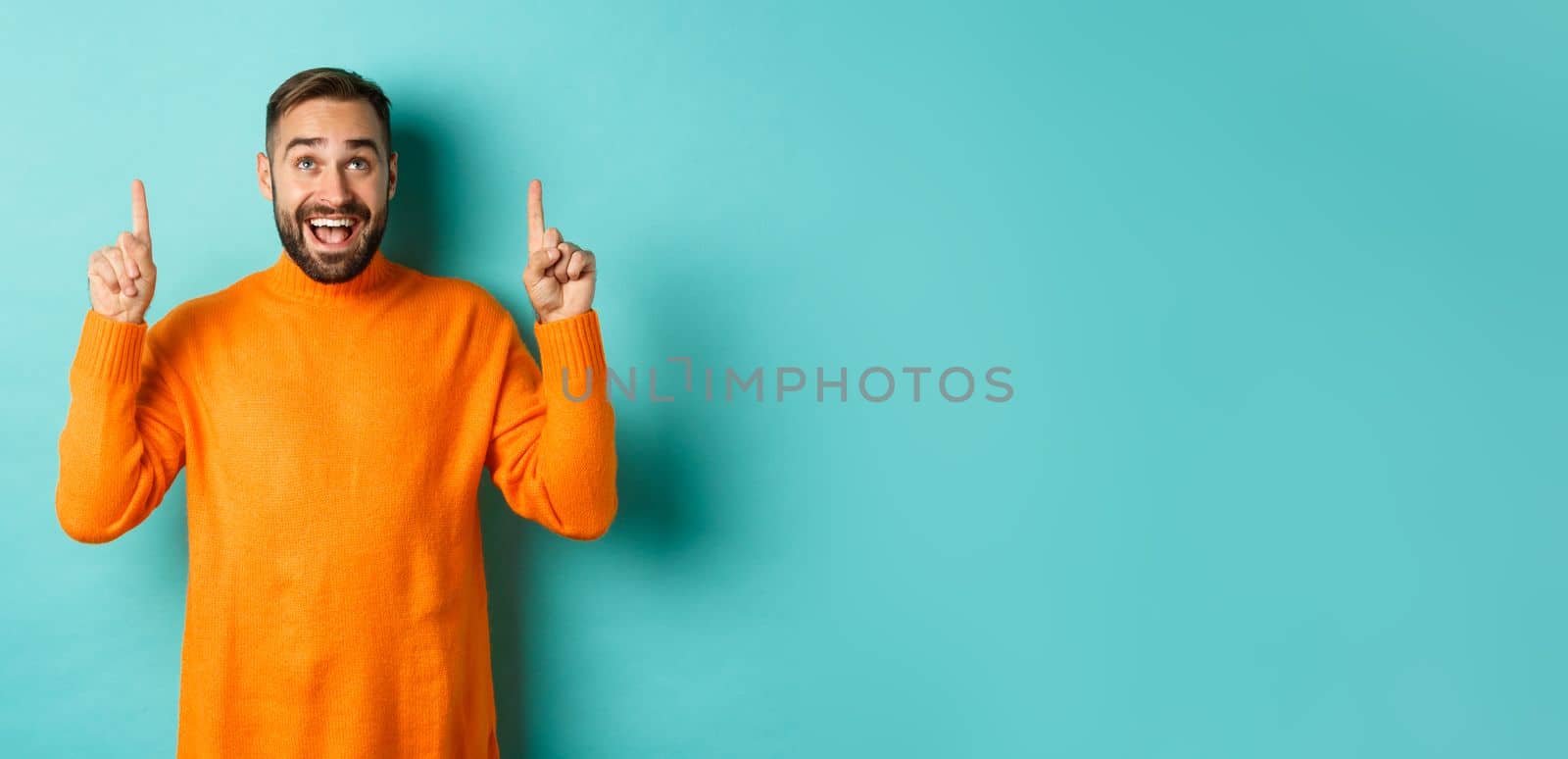 Happy caucasian man showing winter holidays promo offer, pointing fingers up, demonstrate advertisement, standing in sweater against turquoise background.