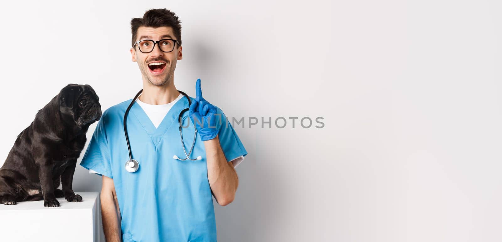 Handsome young doctor at vet clinic pointing finger up and looking amazed, standing near cute black pug dog, white background.