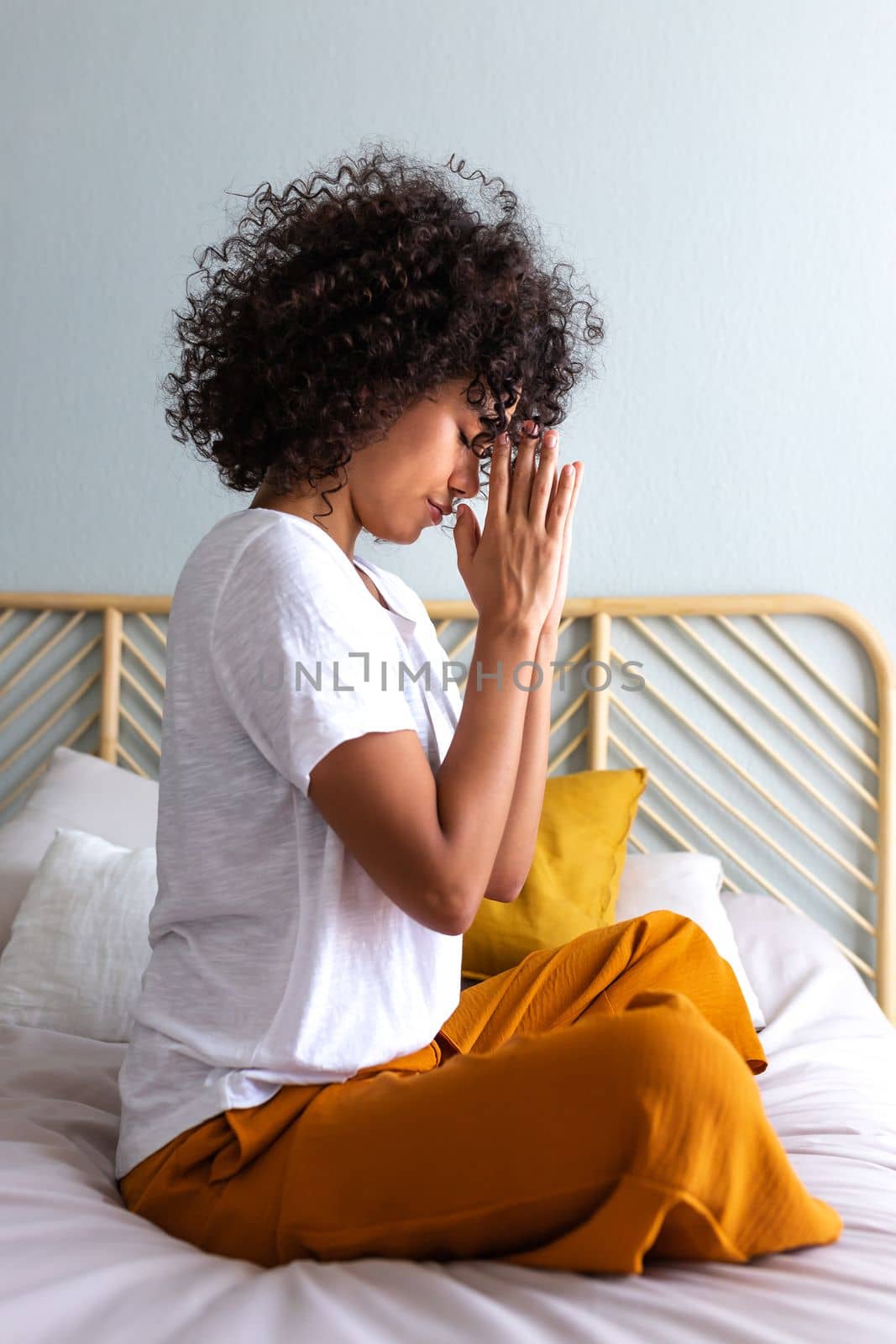 Side view of spiritual young multiracial woman meditating with hands in prayer at home sitting on bed. Vertical image. Spirituality concept.