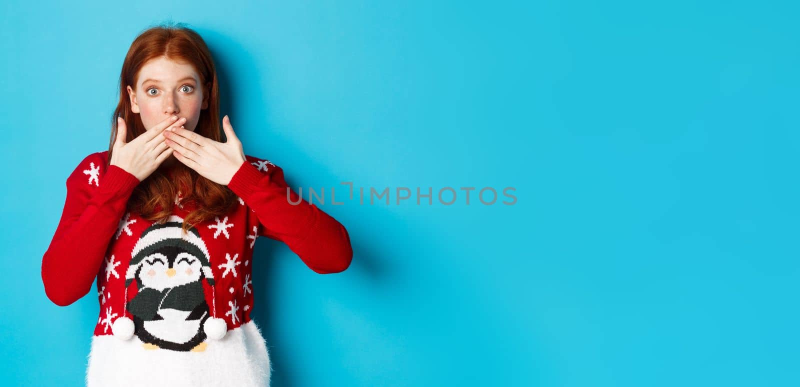 Winter holidays and Christmas Eve concept. Surprised redhead girl in cute sweater, gasping and covering mouth with hands, staring at camera, standing over blue background.