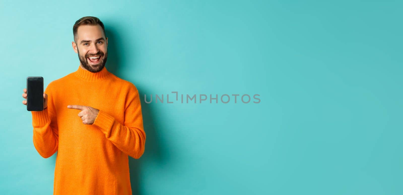 Handsome bearded man in orange sweater, pointing finger at mobile phone screen, showing application smartphone, standing over light blue background.
