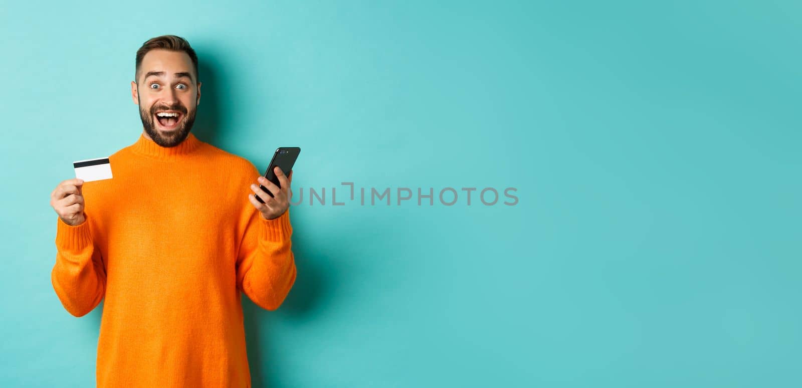 Online shopping. Surprised man holding mobile phone and credit card, paying in internet store, standing over light blue background by Benzoix