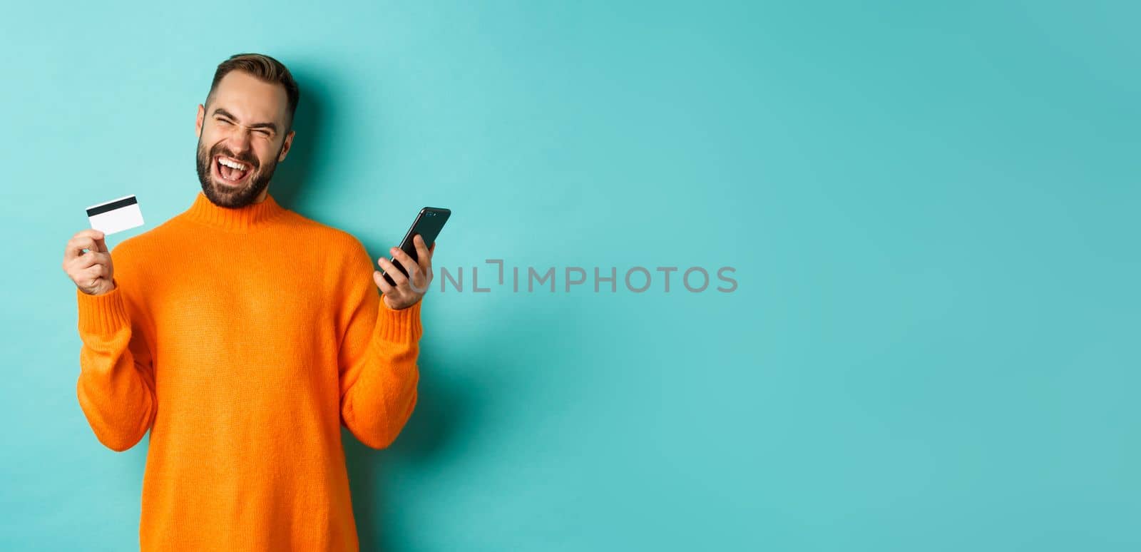 Online shopping. Happy young man using mobile phone and credit card, paying internet, light blue background.