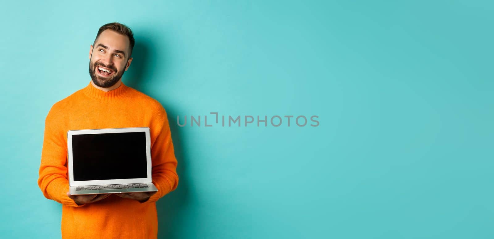 Happy caucasian man thinking and showing laptop screen, smiling pleased while pondering, standing in orange sweater against light blue background.