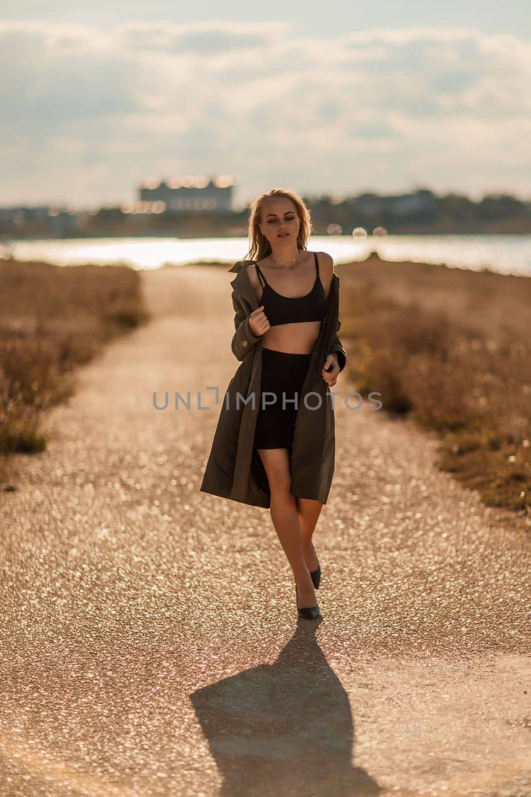 Portrait blonde sea cape. A calm young blonde in an unbuttoned khaki raincoat walks along the seashore, under a raincoat a black skirt and top.