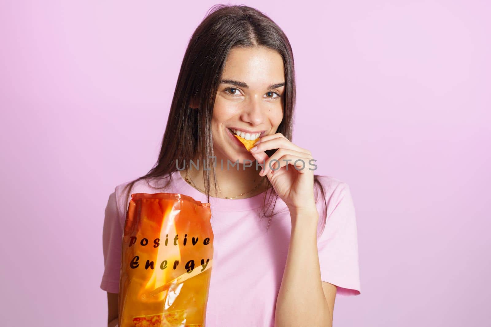 Positive young brunette in colorful t shirt standing on pink background and eating tasty crisps from orange packaging with Positive Energy inscription