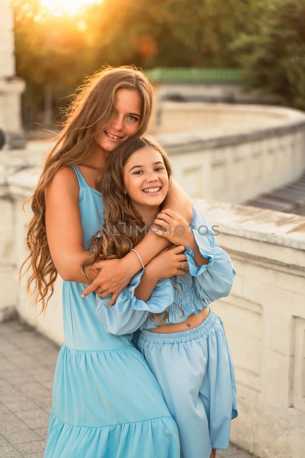 Portrait of mother and daughter in blue dresses with flowing long hair against the backdrop of sunset. The woman hugs and presses the girl to her. They are looking at the camera