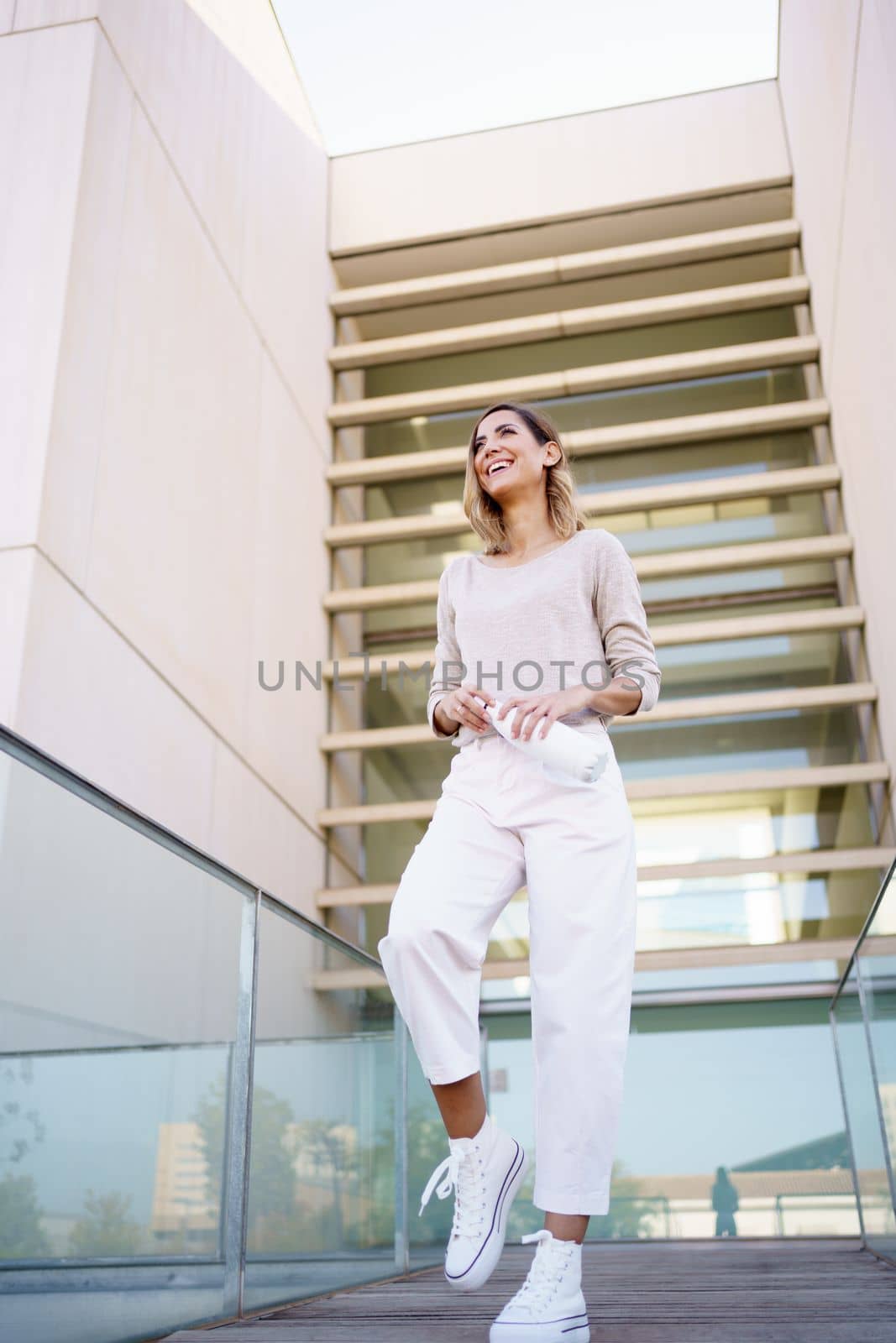 Low angle of cheerful female in stylish outfit, laughing and looking away while standing on path and opening eco friendly bottle of water against modern building on city street