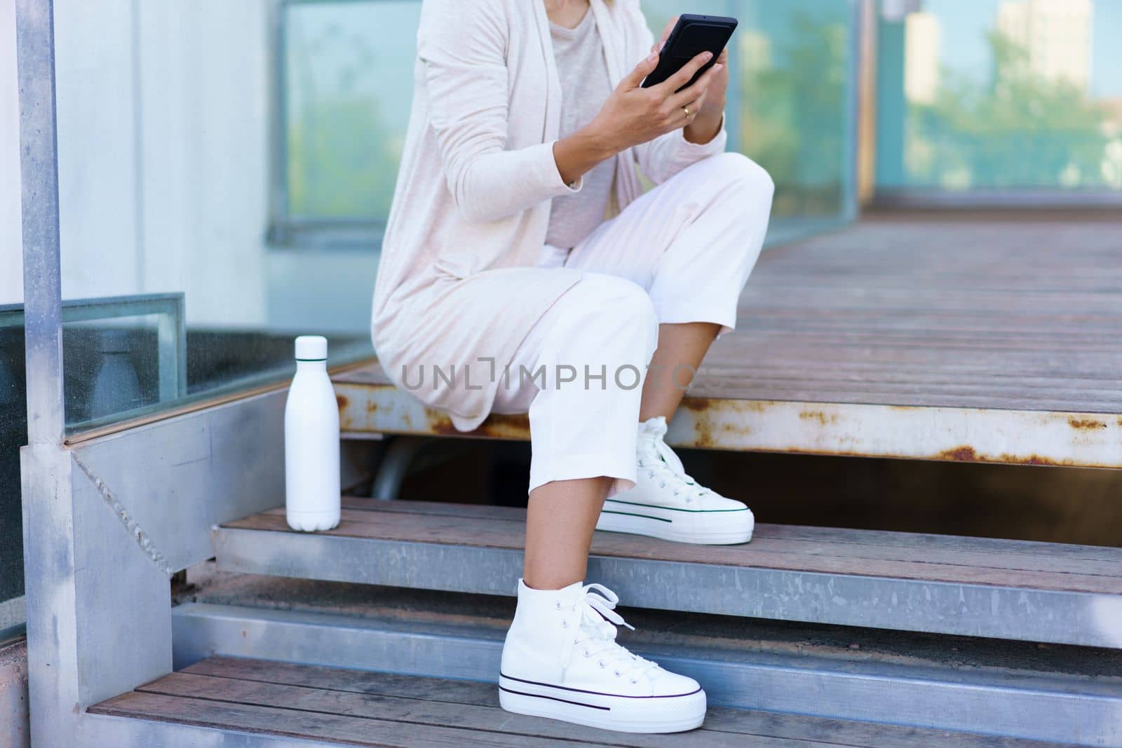 Unrecognizable woman taken a coffee break, sitting on steps near her office building with an eco-friendly ecological metal water bottle. Businesswoman using smartphone.