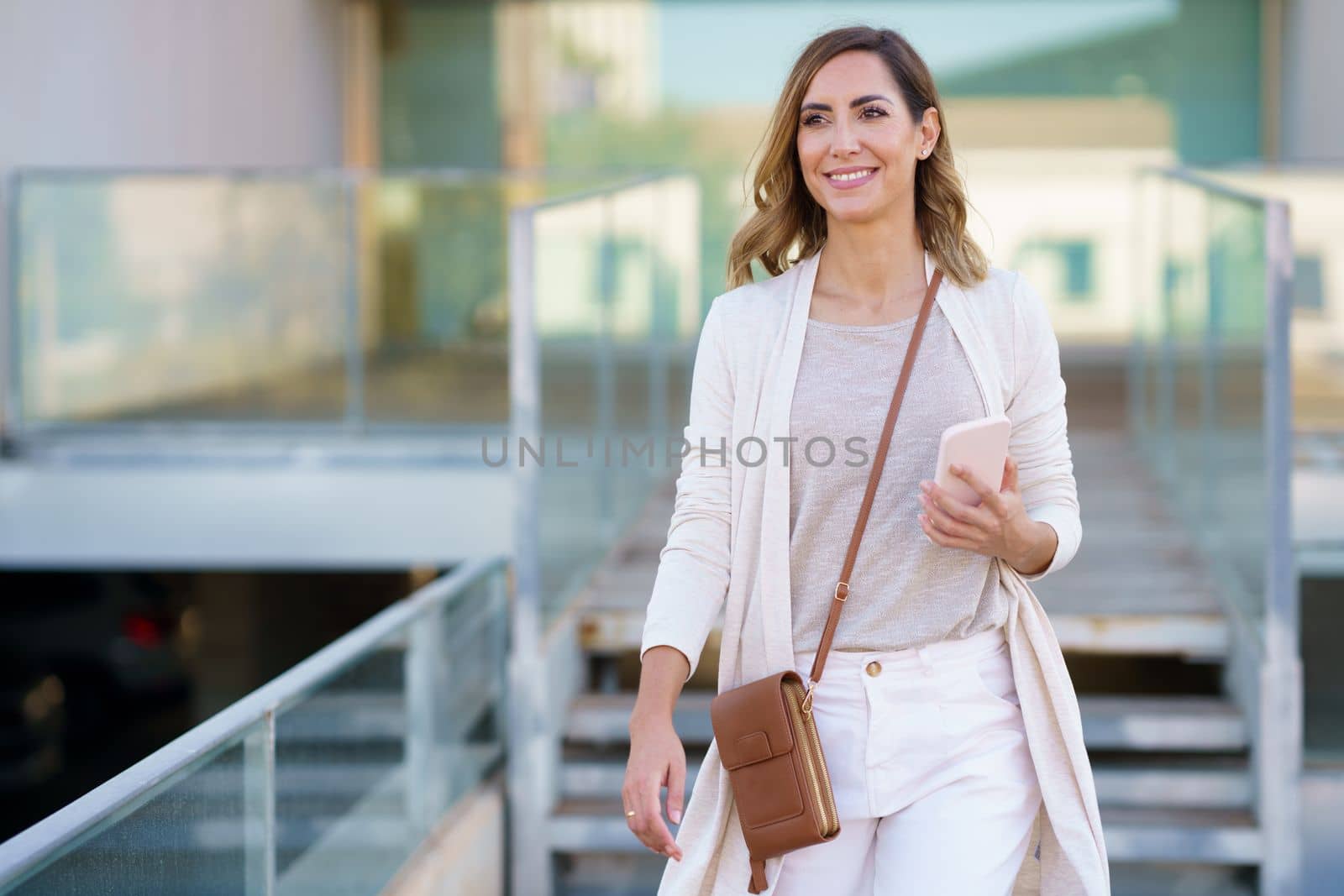 Middle aged female standing near an office building carrying a handbag and a smartphone. Caucasian woman in urban background.