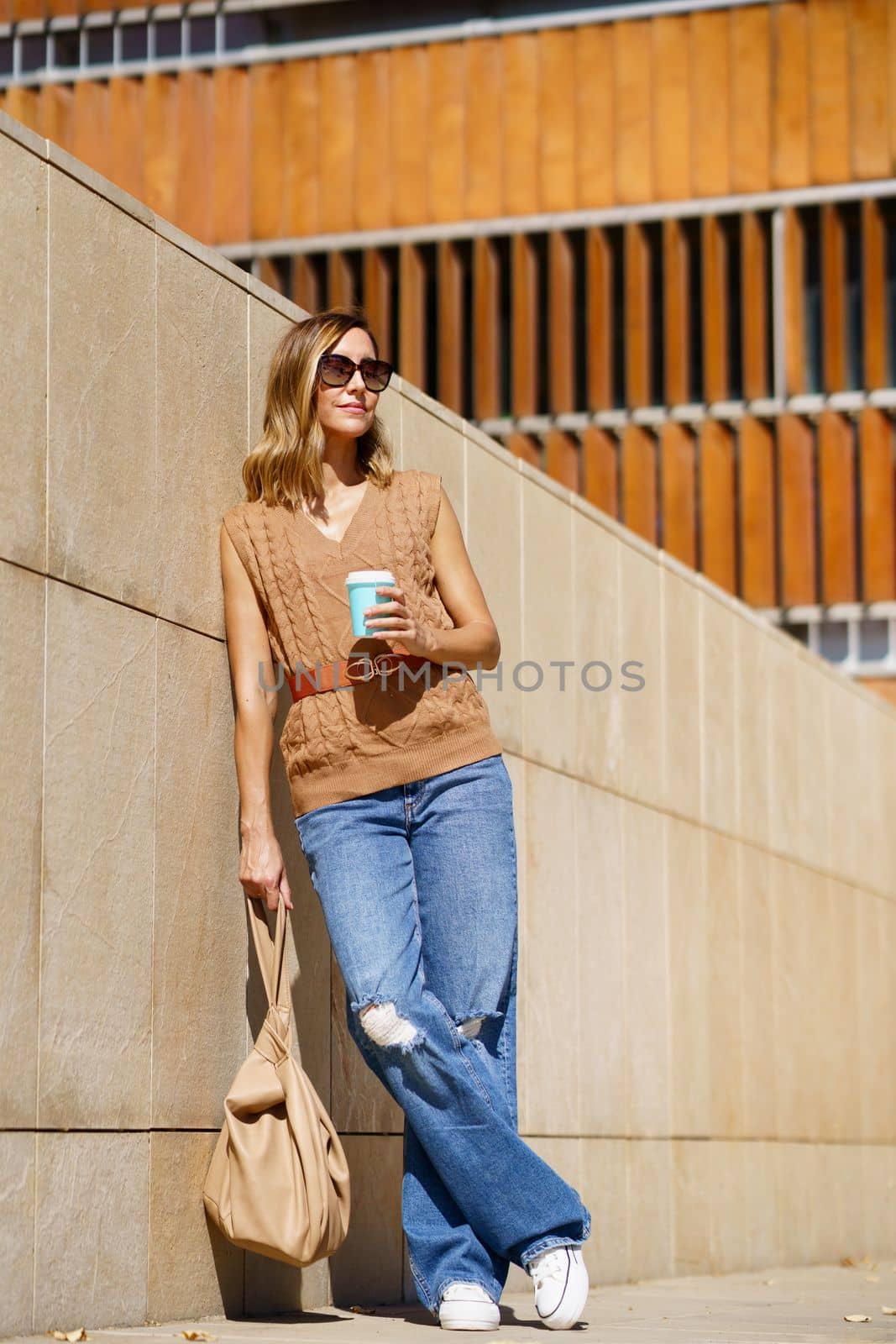 Full body adult female in stylish clothes with bag and coffee to go leaning on wall and looking away on city street