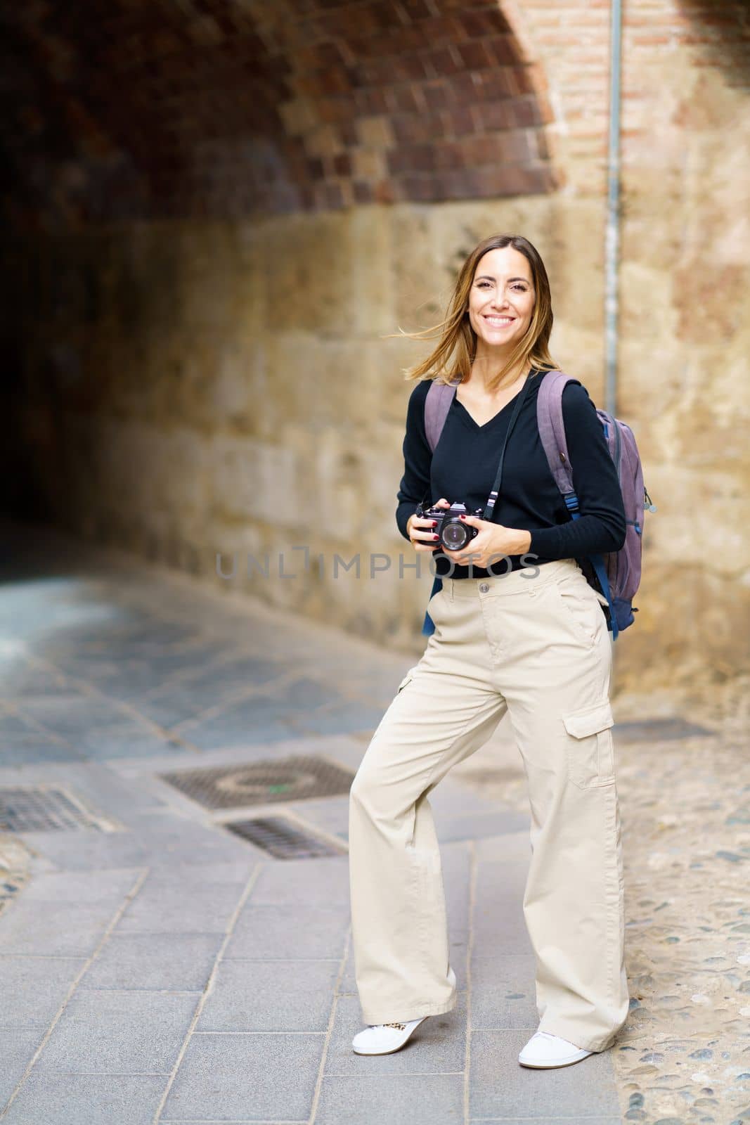 Confident happy young female tourist with long brown hair in casual outfit, and backpack smiling brightly and looking at camera while standing on old town with photo camera