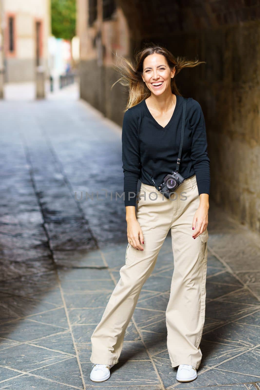 Full length of cheerful young female tourist with photo camera in casual outfit, laughing and looking at camera while standing on narrow street in old town
