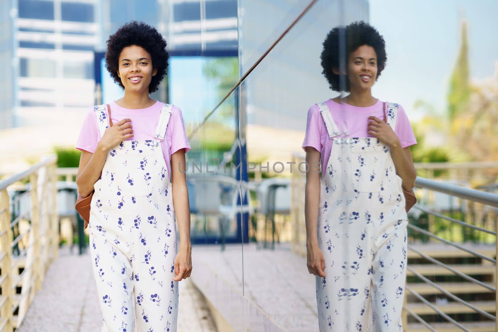 Happy African American young female with Afro hairstyle in overall looking away with smile while standing near glass building in city