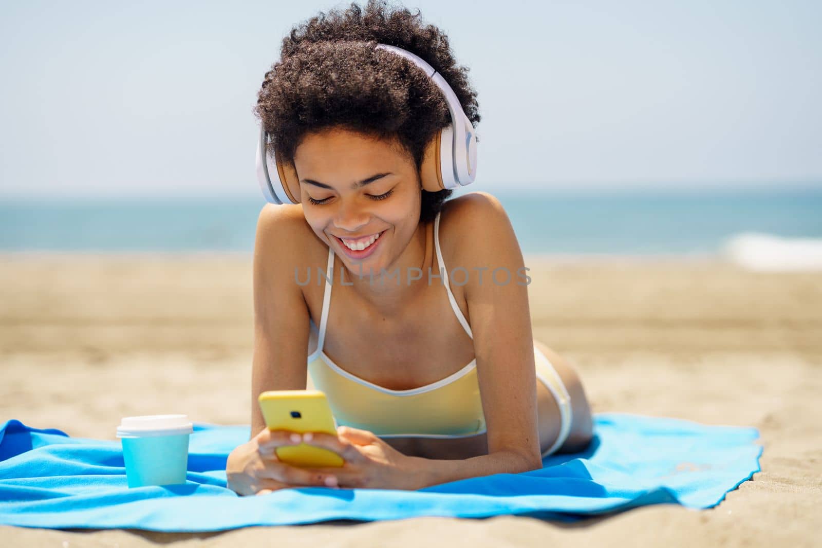 Cheerful young African American woman with curly dark hair, in bikini smiling and using mobile phone while, listening to music in headphones and lying on sandy beach on sunny day
