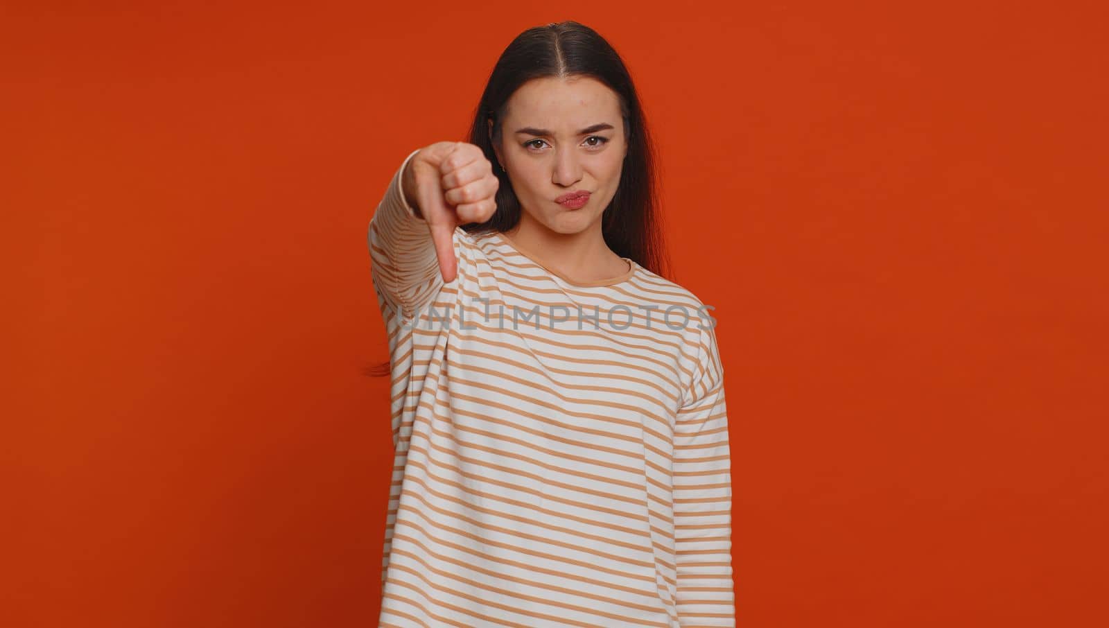 Dislike. Upset unhappy pretty woman in pullover showing thumbs down sign gesture, expressing discontent, disapproval, dissatisfied, dislike. Young adult girl. Indoor studio shot on red background