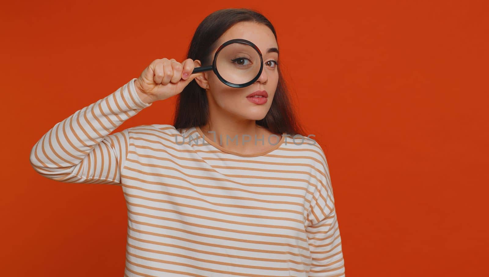 Investigator researcher scientist woman holding magnifying glass near face, looking into camera with big zoomed funny eyes, searching, analysing. Young pretty girl isolated on red studio background