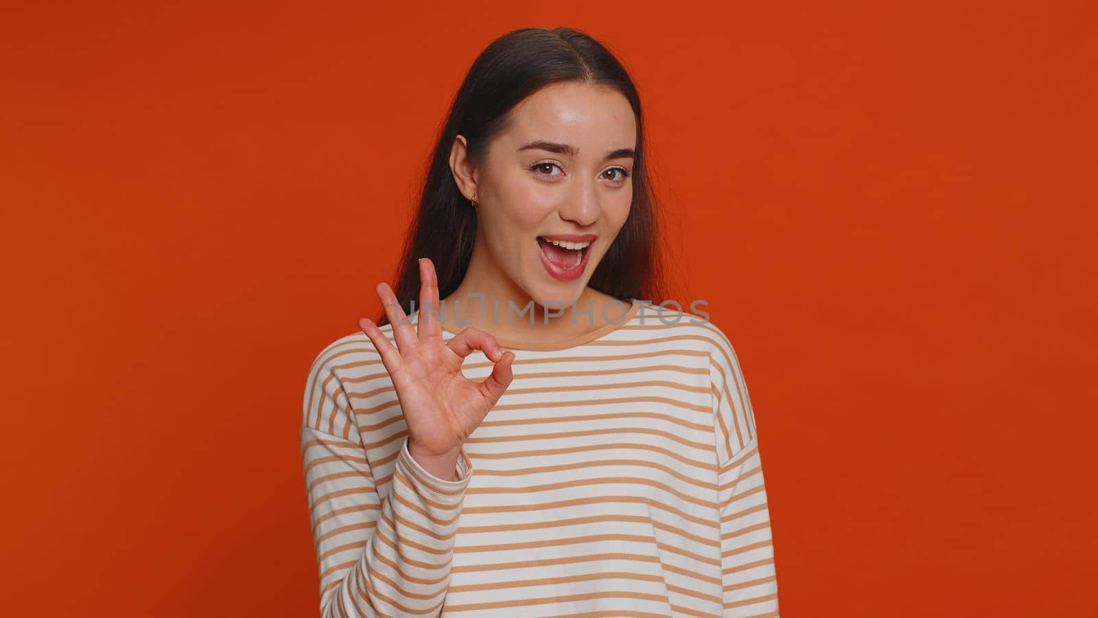 Ok. Joyful happy pretty woman in pullover looking approvingly at camera showing approve feedback gesture like sign positive something good. Young girl on red studio background. People sincere emotion
