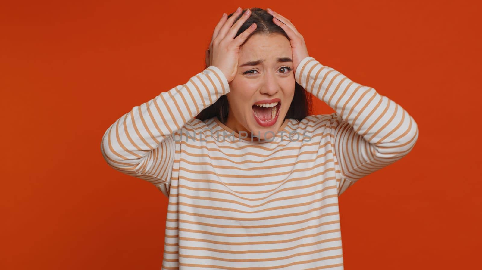 Young woman in pullover screams from stress tension problems feels horror hopelessness fear surprise shock expresses gestures rage. Pretty brunette girl indoors isolated alone on studio red background