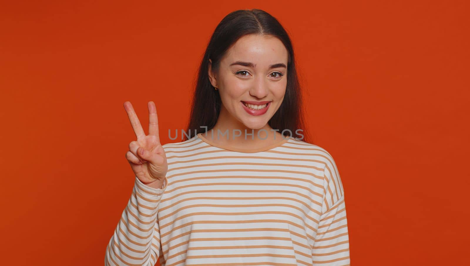 Hipster pretty woman in pullover showing victory v sign, hoping for success and win, doing peace gesture, smiling with kind optimistic expression. Young adult girl isolated on red studio background