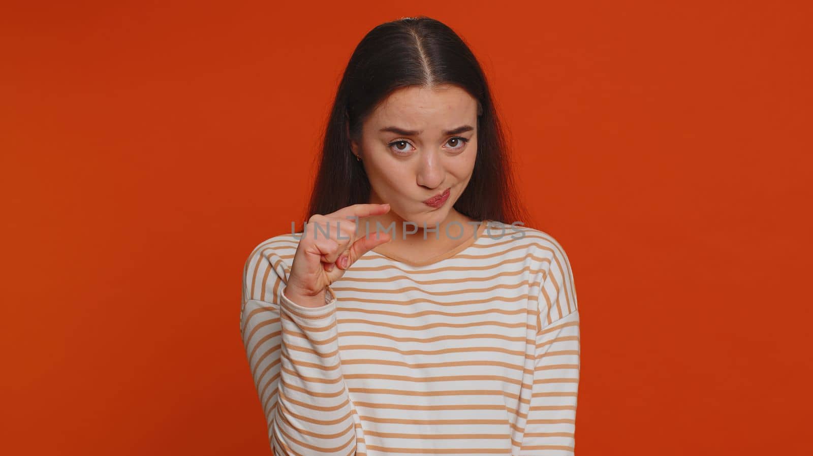 Need some more, please give me. Lovely woman showing a little bit gesture with sceptic smile, showing minimum sign, measuring small size. Young adult girl isolated alone on red studio background