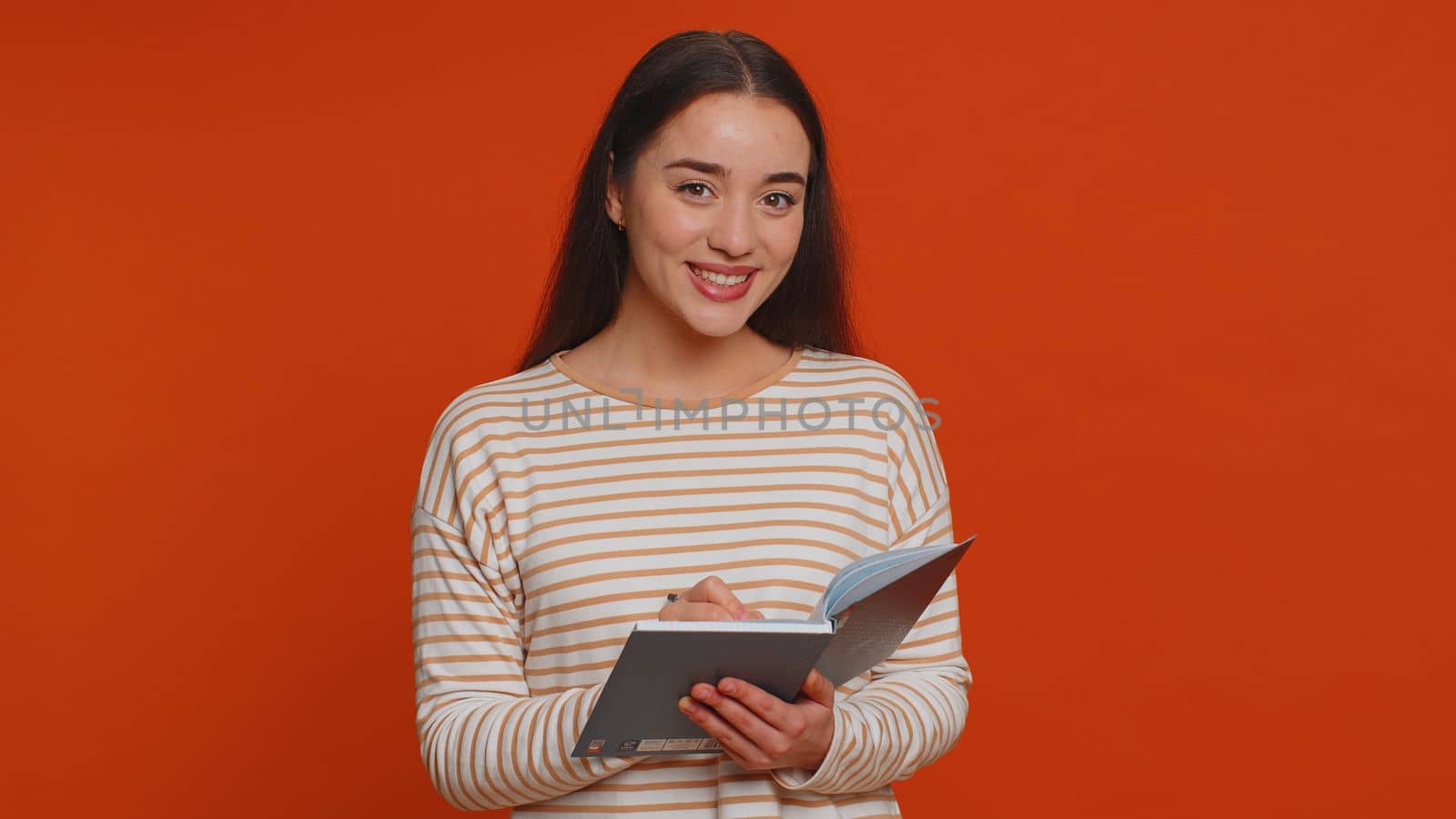 Thoughtful journalist pretty young woman in pullover making notes, writing down thoughts with pen into notepad notebook diary, to do list, good idea. Millennial girl isolated on red studio background