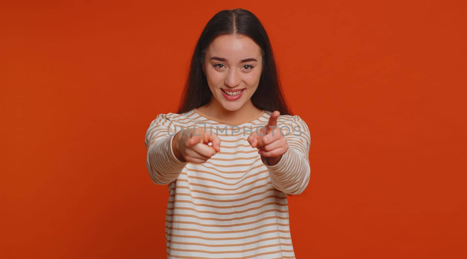 Hey you. I choose you. Young woman in pullover smiling excitedly and pointing finger to camera, choosing lucky winner, indicating to awesome you. Pretty girl indoors isolated on studio red background