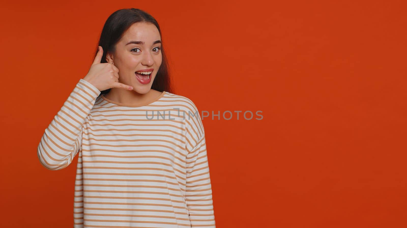 Call us, here is contact number. Young woman looking at camera doing phone gesture like says hey you call me back, conversation. Caucasian girl showing advertising area isolated on red background