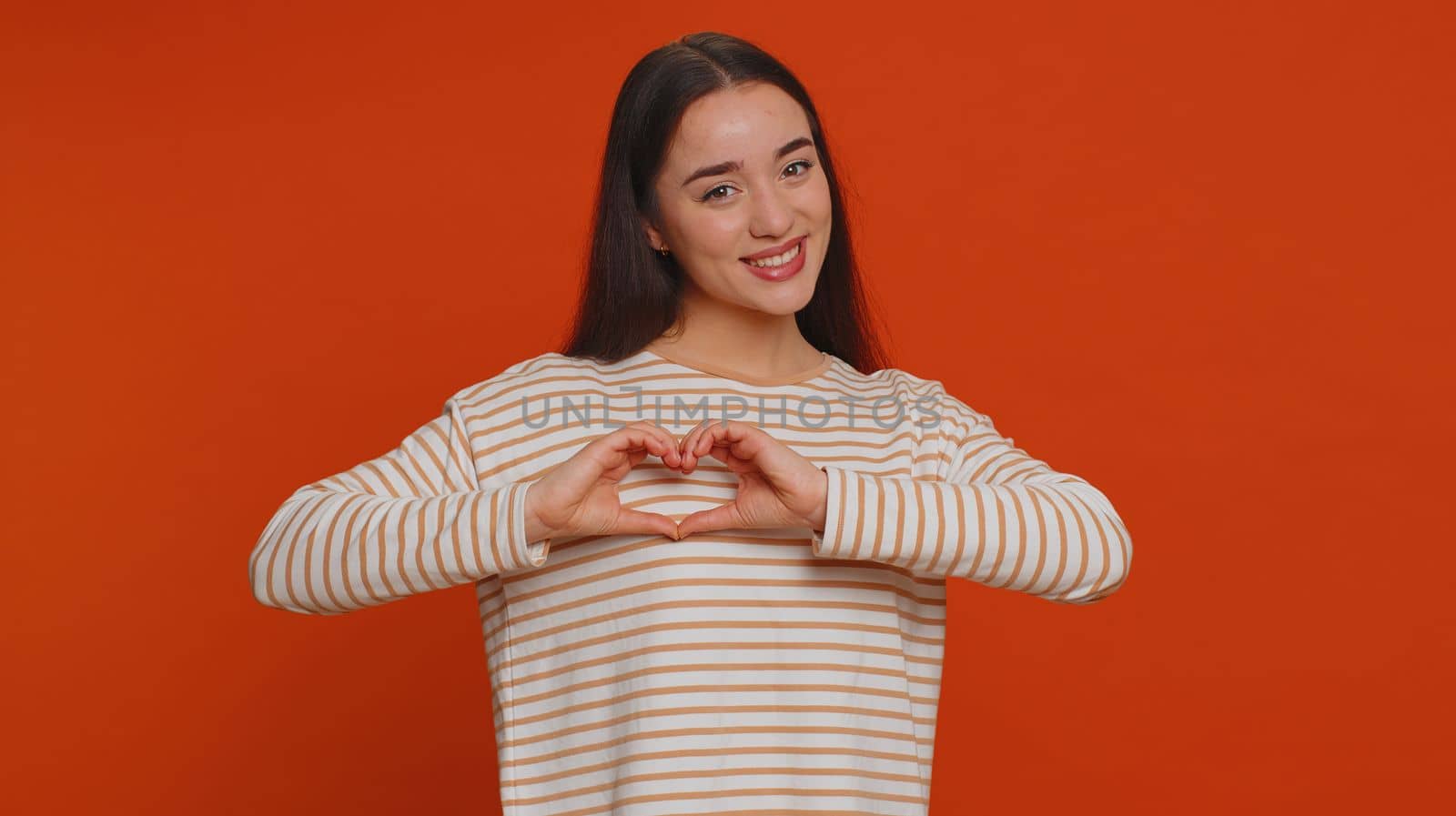 Woman in love. Smiling pretty woman 20s in pullover use makes heart gesture demonstrates love sign expresses good feelings and sympathy. Young one adult girl isolated alone on red studio background