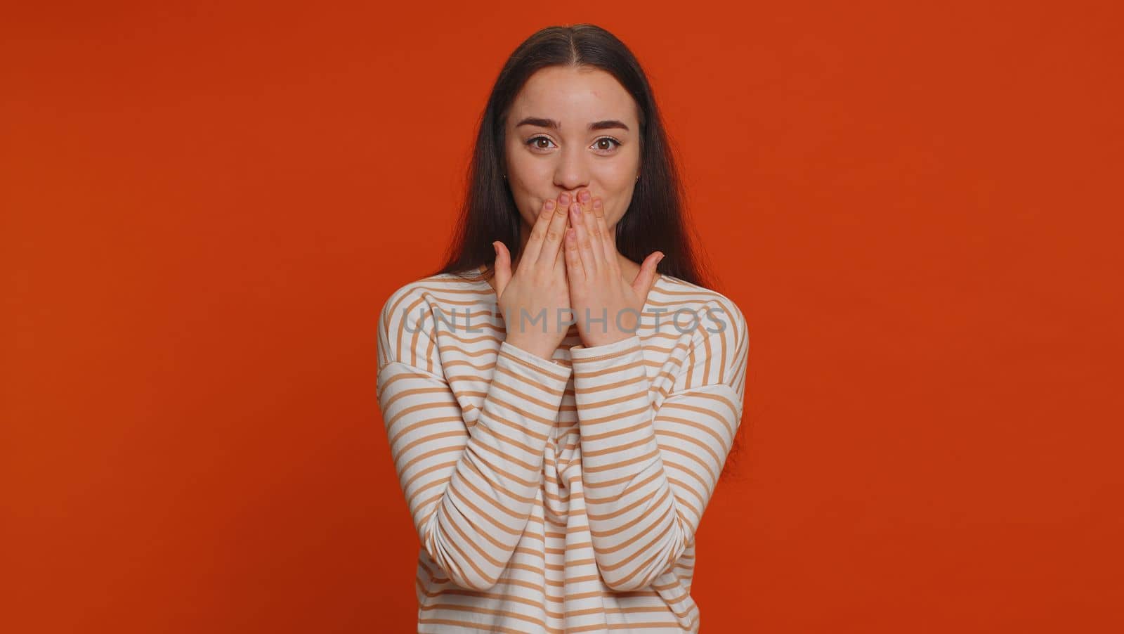 I will not say anyone. Frightened woman closing her mouth with hand, looking intimidated scared at camera, gestures no, refusing to tell terrible secret, unbelievable truth. Girl on red background