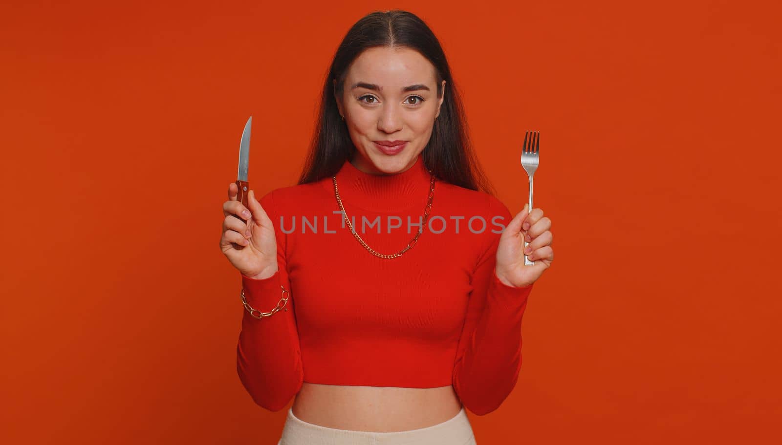 Ready to eat. Hungry young woman in crop top waiting for serving dinner dishes with cutlery, will appreciate delicious restaurant meal menu. Girl holding fork and knife on red studio background