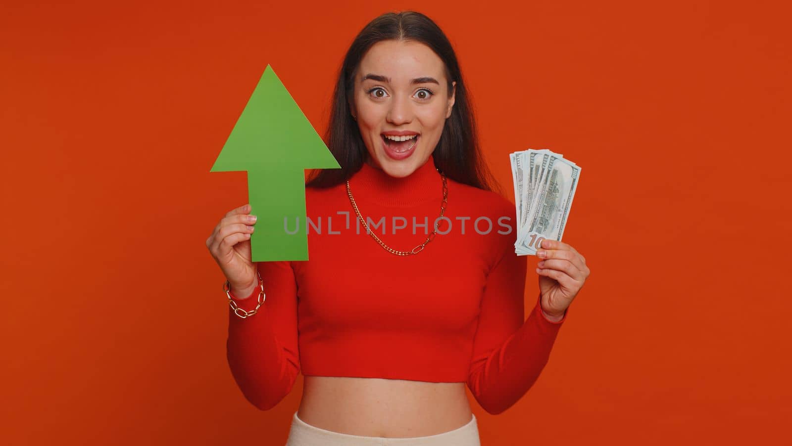 Happy pretty woman winner holding arrow sign pointing up, career growth and money dollar exchange increase. Young adult girl showing up indicating successful development progress on red background