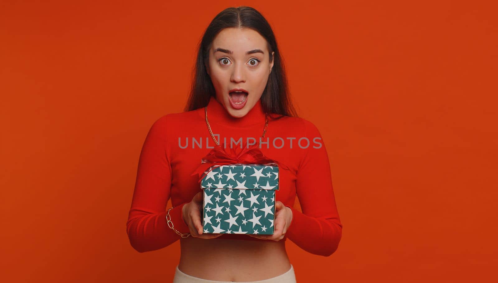 Happy pretty woman opening gift box, unwrapping birthday surprise and expressing great happiness amazement, satisfied with best present bonus. Young adult lovely girl isolated on red studio background