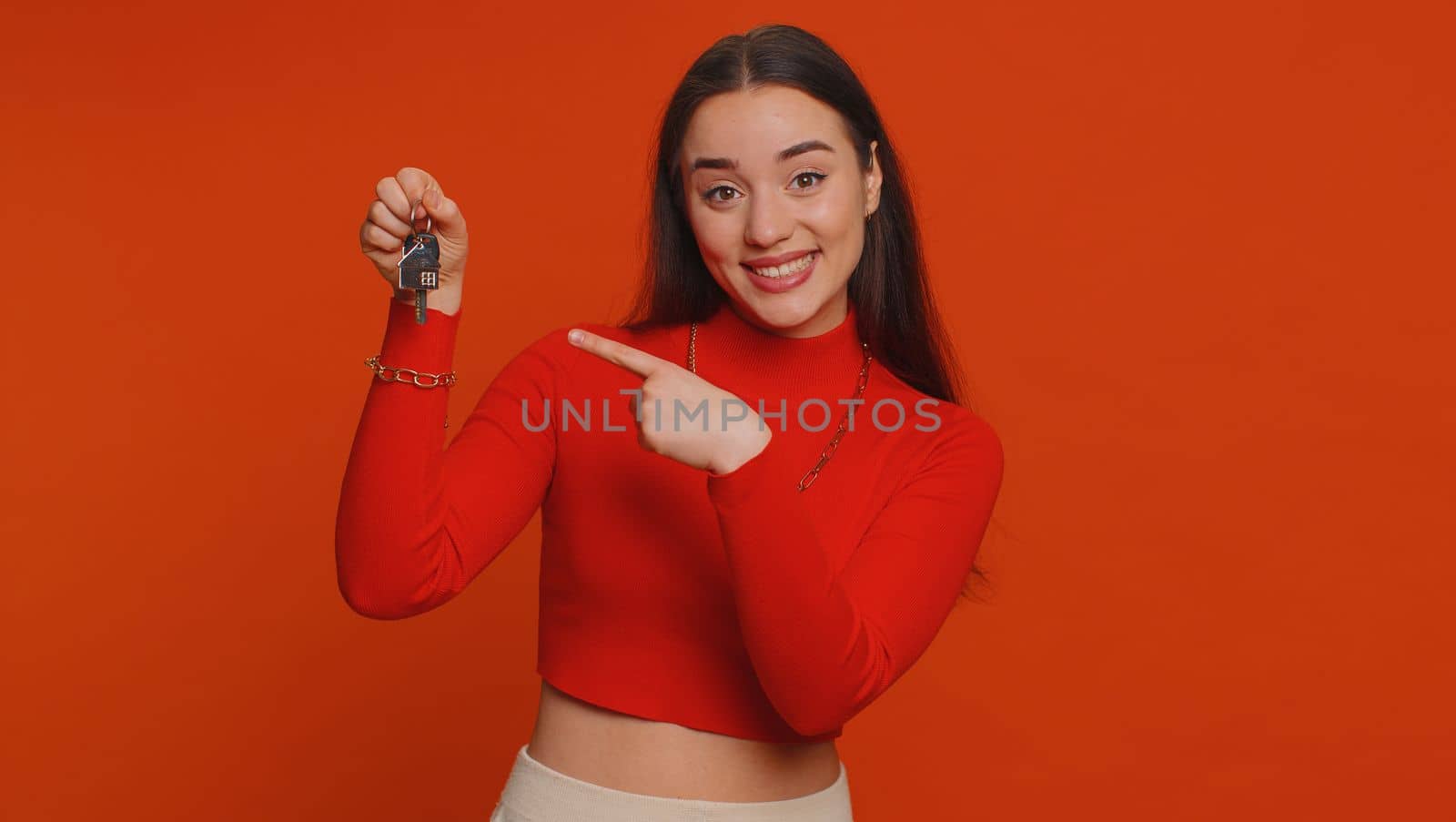 Pretty young woman in crop top real estate agent lifting hand showing the keys of new home house apartment, buying, renting property, mortgage loan. Millennial girl isolated on red studio background