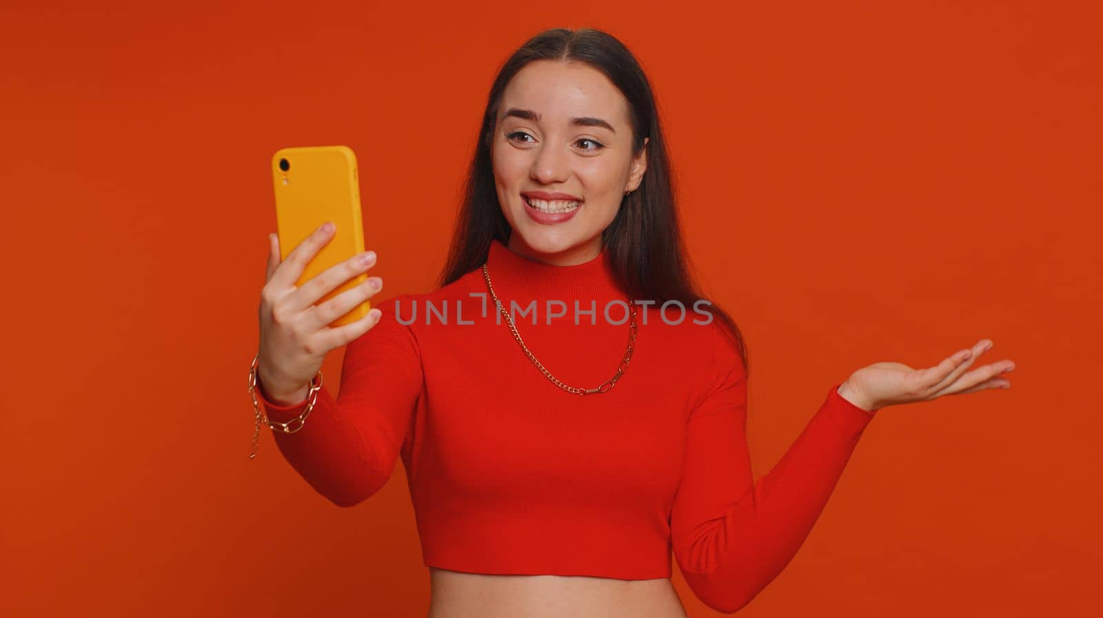Pretty young woman influencer blogger taking selfie on smartphone, communicating video call online, recording new social media vlog story for subscribers. Girl isolated on red studio background