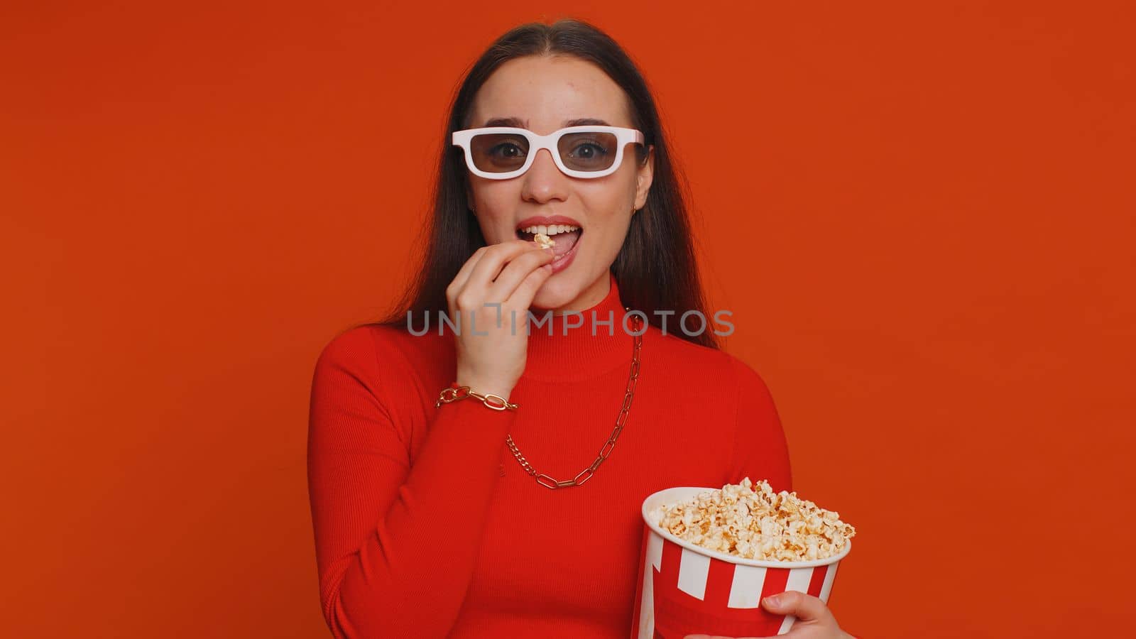 Excited pretty woman in 3D glasses eating popcorn and watching interesting TV home serial, sport game, film, online social media movie content. Girl enjoying domestic entertainment on red background
