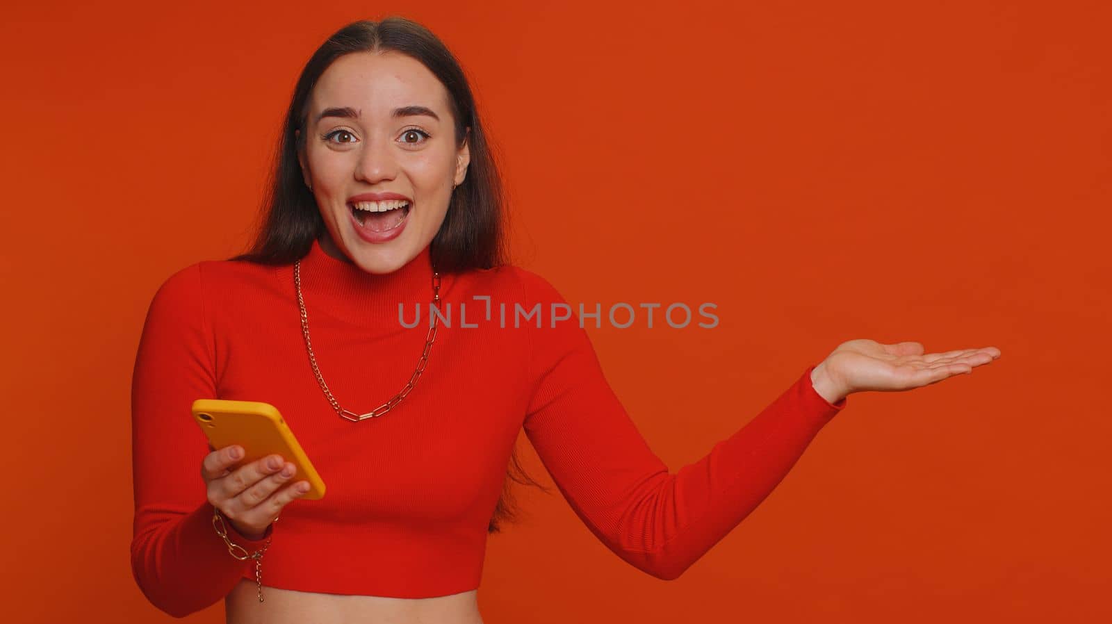 Woman using smartphone pointing empty place, advertising area for commercial text, copy space for goods application promotion social networks positive feedback. Girl isolated on red studio background