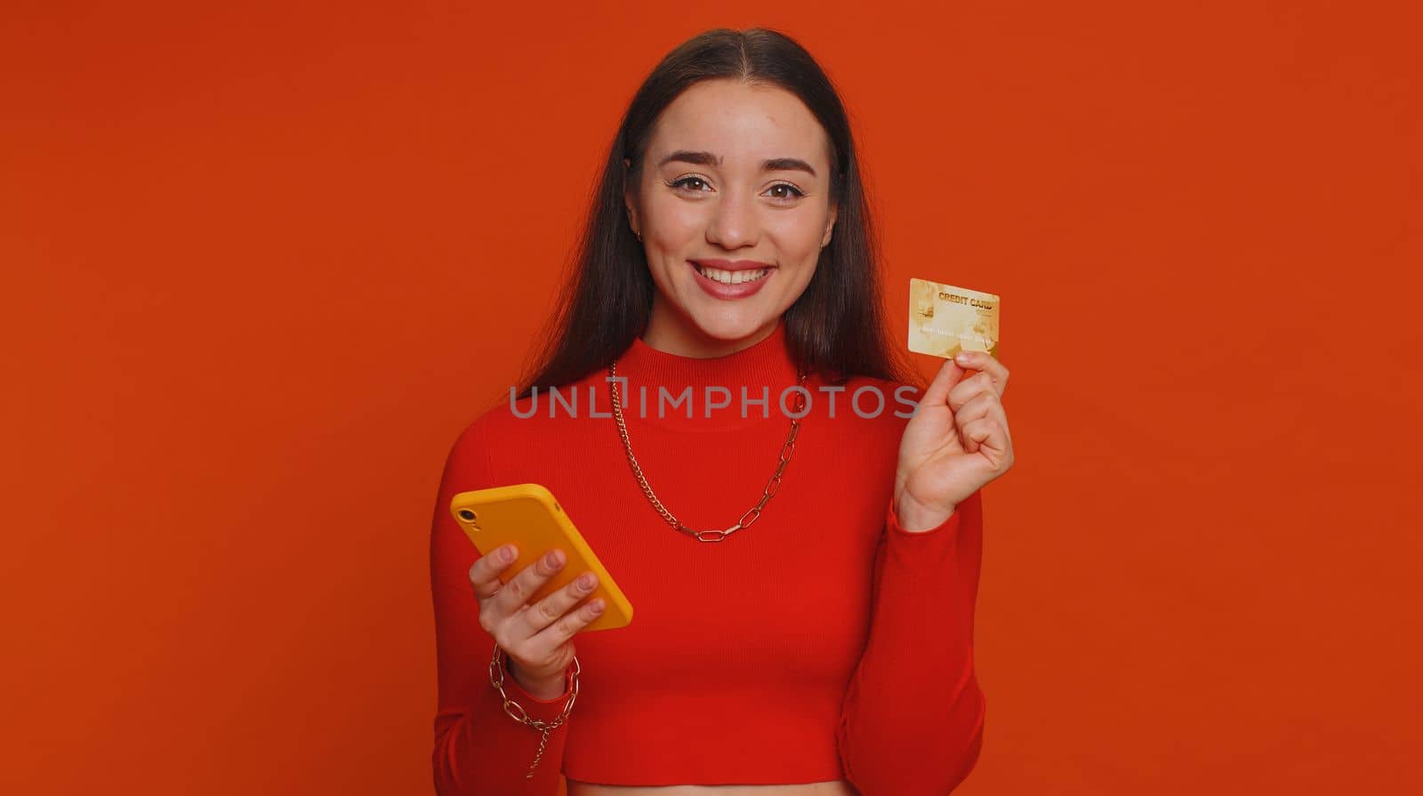 Sincere pretty woman customer using credit bank card and smartphone while transferring money, purchases online shopping, payment. Finance and internet. Young girl isolated alone on red background