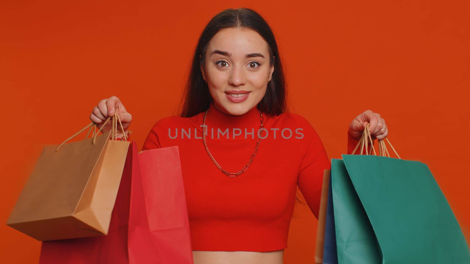 Young woman in crop top showing shopping bags, advertising discounts, smiling looking amazed with low prices, shopping on Black Friday holidays. Girl indoors isolated alone on studio red background