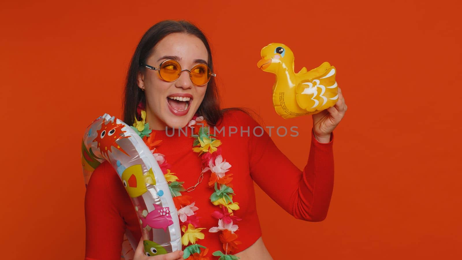 Traveler tourist young woman celebrating, dancing fooling with swimming inflatable duck toy. Concept summer vacation, journey, trip to seaside. Lovely girl in sunglasses on red studio background
