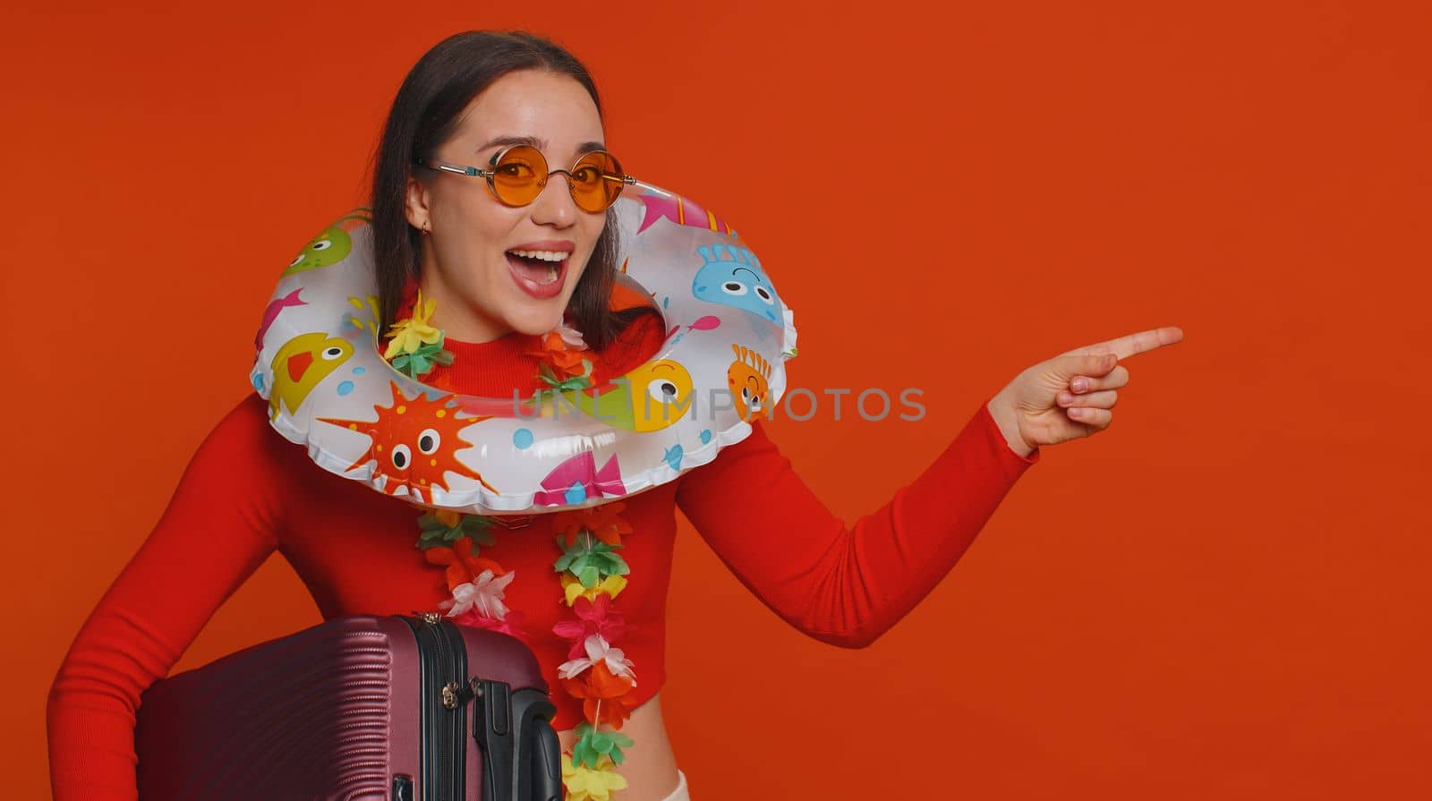 Cheerful happy tourist woman with luggage bag, swimming ring pointing empty place, advertising area for commercial text, copy space for goods services banner promotion. Vacation, summer trip to sea