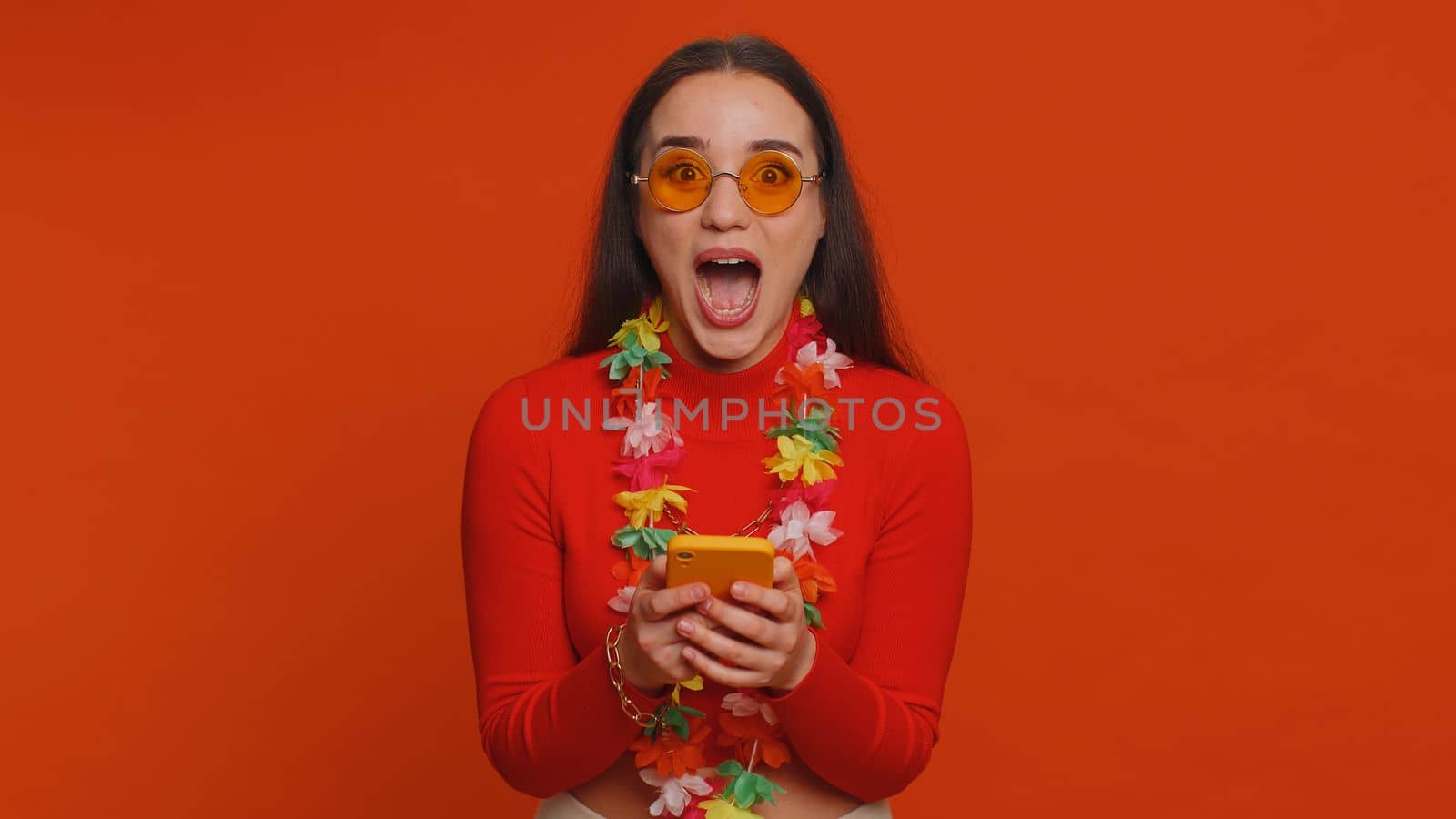Cheerful tourist woman with hawaiian lei in sunglasses using smartphone celebrating winning holiday resort vacation tickets pass. Tourism, summer trip to sea. Happy pretty girl on red wall background