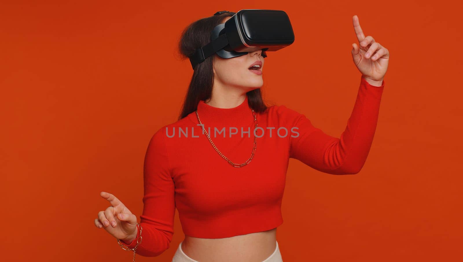 Lovely woman using headset helmet to play simulation game app online. Watching virtual reality 3D 360 video content. One girl in VR goggles isolated on studio red background. Futuristic technology