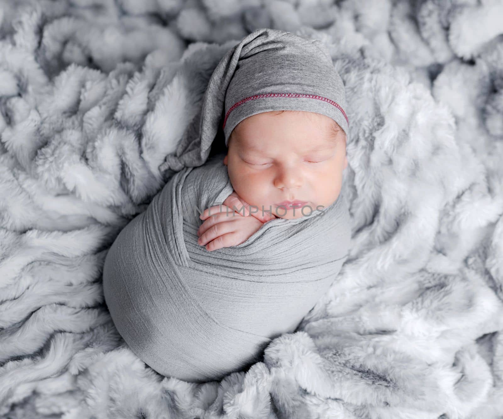 Cute newborn baby boy swaddled in grey fabric sleeping on fur blanket. Adorable infant child kid napping studio portrait