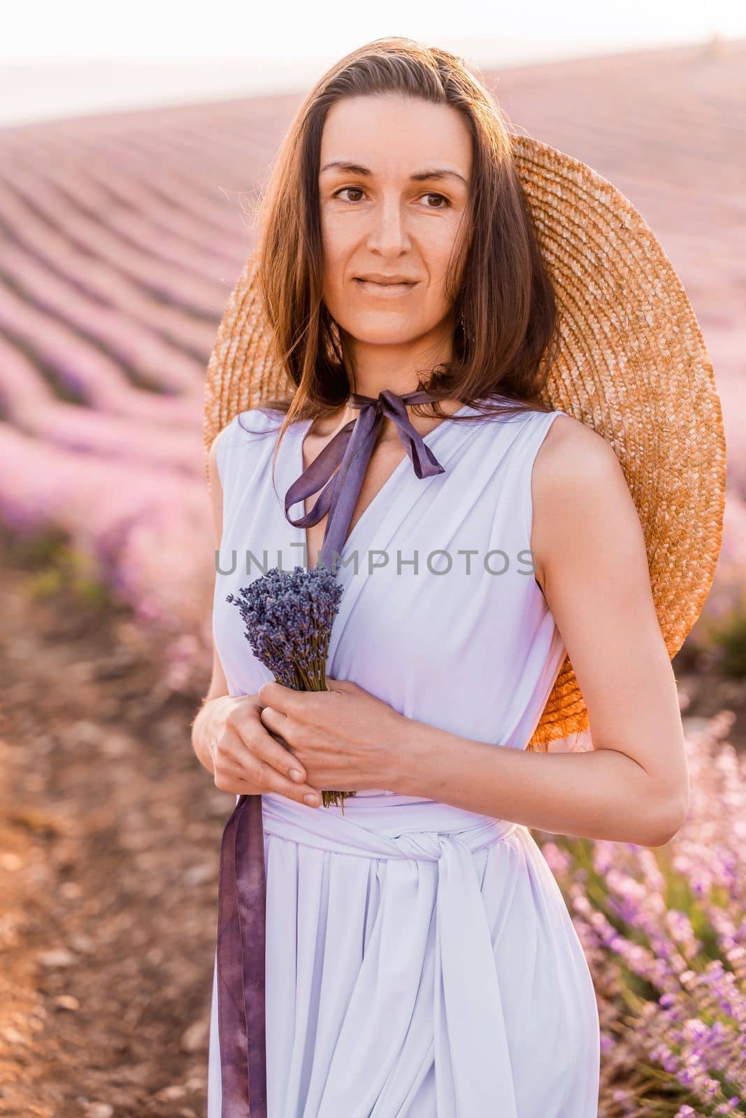 Woman lavender field sunset. Romantic woman walks through the lavender fields. illuminated by sunset sunlight. She is dressed in a white dress with a hat