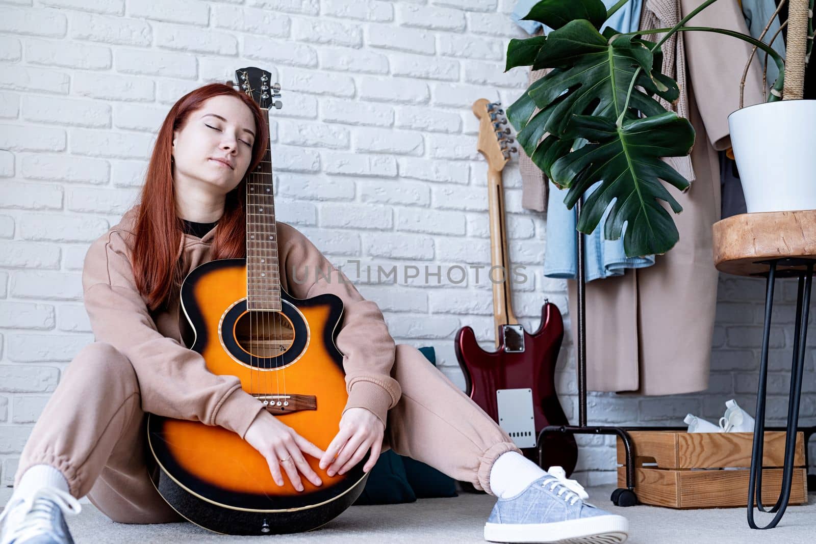 young caucasian red-haired woman sitting on rug and playing acoustic guitar at home